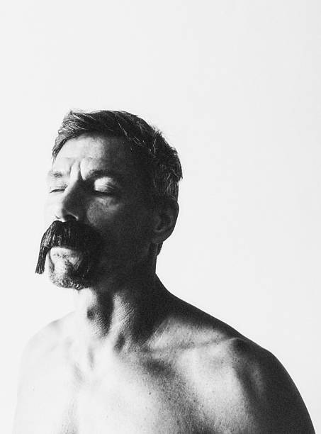 A middle-aged man with closed eyes, bare-shouldered, wearing an exaggerated fake mustache. The bright, nearly overexposed light gives the scene a fragile, dreamlike quality between self-exploration and transformation.