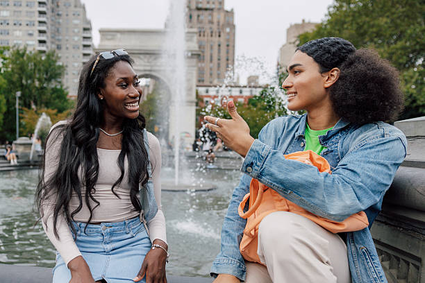 Two friends stroll in Washington Square filled with trees and greenery, engaging in a cheerful conversation sitting by the fountain.