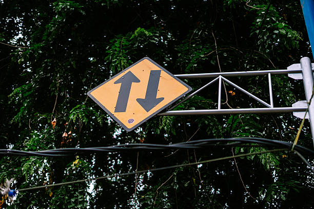 A bright yellow diamond-shaped road sign with black arrows pointing up and down, indicating two-way traffic, set against a background of lush green foliage and dark power lines.