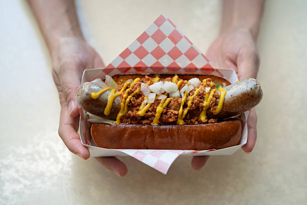Hands holding gourmet hot dog with sausage, minced meat, onion, and mustard