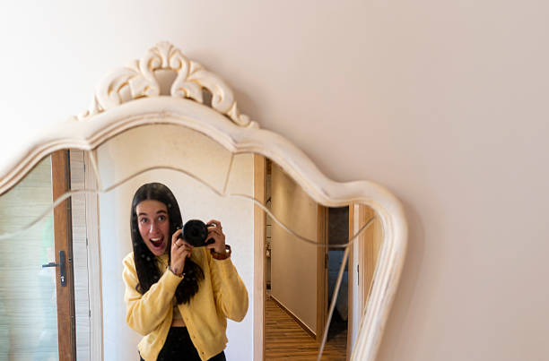 Young female photographer capturing herself in a vintage-style mirror with an excited expression, holding a professional camera, reflecting an interior hallway