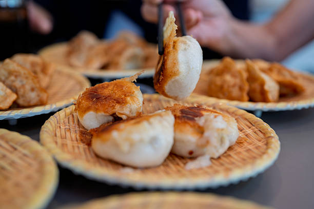 Close-up of freshly pan-fried pork buns with crispy golden bottoms served on plates in a Chinese restaurant. A hand holding chopsticks picks up one dumpling, showing texture and flavor of Asian street food.