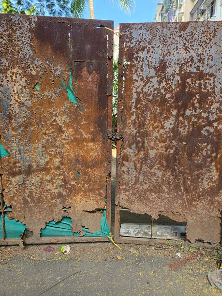 Close up view of heavily rusted metal sheets used as a temporary construction fence. Corroded surface texture and peeling paint reveal decay, with scattered dry leaves and debris on the ground showing urban wear and neglect.