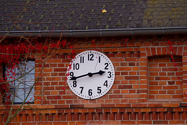 A brick wall with a round clock mounted on it, part of a window and branches with red leaves are also visible.