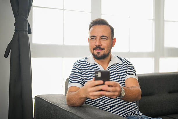 istock Smiling man sitting on a sofa and using smartphone at home with natural light background 2241979835