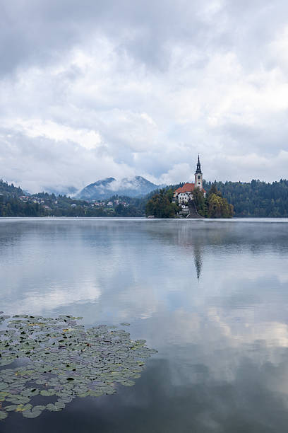 Calm lake with mist rising and a church on a small island surrounded by forested hills. Scenic water reflection and cloudy sky create a peaceful nature landscape. Tranquil outdoor scene.