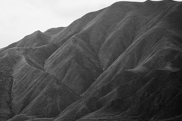 Mesmerizing black and white abstract landscape photograph showcasing the intricate, rhythmic contours of weathered hills and shadowed valleys, with subtle mist enhancing depth and geological drama. The high-contrast composition highlights natural erosion patterns, textured ridges, and serene isolation, creating a timeless study in form and texture. Ideal for themes of abstract earth science, minimalist serenity, environmental artistry, or topographic inspiration. High-resolution, royalty-free stock image perfect for editorial features, interior design, conceptual photography, book illustrations, or digital wallpapers.