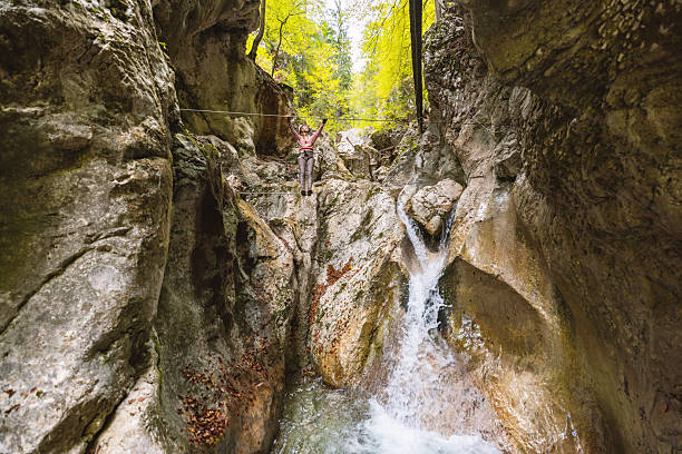 Young woman balancing on rope bridge above rocky canyon with waterfall. Adventurous adult hiking in forested mountain gorge. Outdoor activity in nature with autumn foliage.