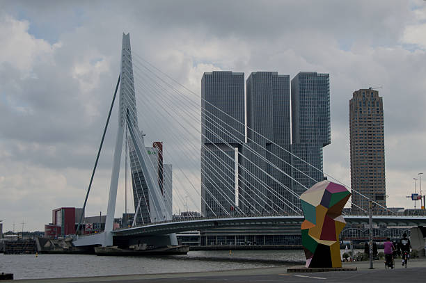 View of the Erasmus Bridge and the modern skyline of Rotterdam, Netherlands, featuring the De Rotterdam building complex and a colorful geometric sculpture in the foreground. The cloudy sky adds a soft, moody light ideal for illustrating architecture, city life, or travel themes.