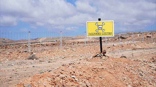 Yellow danger sign with skull and crossbones warning about landmines in a desolate, fenced area after a war conflict, symbolizing the ongoing danger and clearance efforts