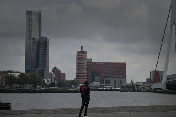 A lone figure stands near the Erasmus Bridge looking at the skyline of Rotterdam, the Netherlands. The overcast weather and modern architecture create a cinematic urban mood, blending introspection with the rhythm of city life. Perfect for creative projects about travel, solitude, architecture, or urban identity.September 2024