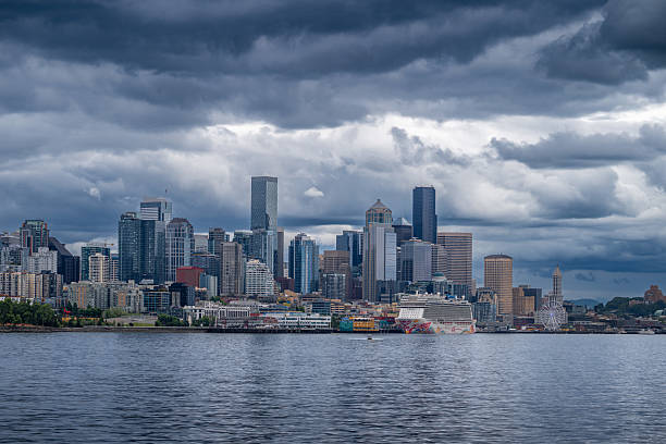 A dramatic view of the Seattle skyline under heavy storm clouds, with the waterfront and a cruise ship visible in the foreground. The dark sky contrasts with the city’s modern architecture, capturing the moody charm of the Pacific Northwest.
