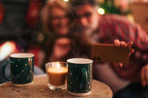 Older adult couple blurred in the background taking a smartphone selfie while cozy mugs and a lit candle glow in the foreground, celebrating christmas at home together