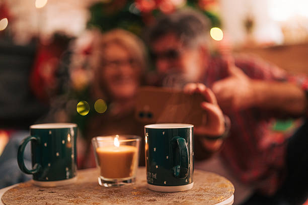 Unfocused mature adult couple taking a selfie with a smartphone by a lit candle and two steaming mugs on a cozy wooden table, celebrating christmas holidays at home indoors