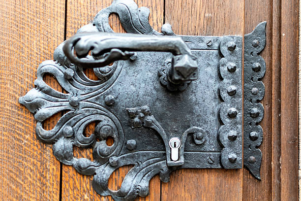 Close-up image of an elaborate, antique black wrought iron door handle and lock plate, mounted on a rustic wooden door with visible grain
