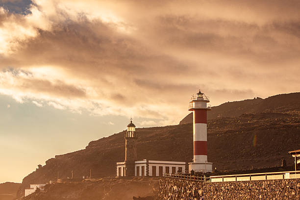 Close-up of the two historic lighthouses set against a volcanic hill, bathed in the warm, intense light of a sunset with dramatic, textured clouds and a golden atmosphere.