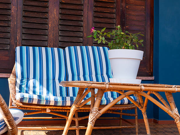cozy balcony features wicker loveseat with blue striped cushions and round table, paired with potted plant. Warm sunlight enhances inviting atmosphere