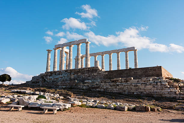 Wide-angle view of the Temple of Poseidon with marble columns, stone steps, and benches under bright blue sky at Cape Sounion, Greece