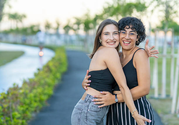 A joyful portrait of two women embracing each other outdoors, both smiling warmly at the camera.
