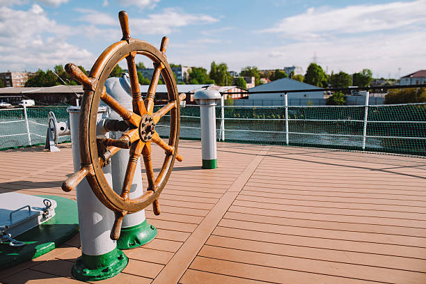 Wooden steering wheel on open ship deck under sunlight with city harbor view in background, nautical navigation, sea transport
