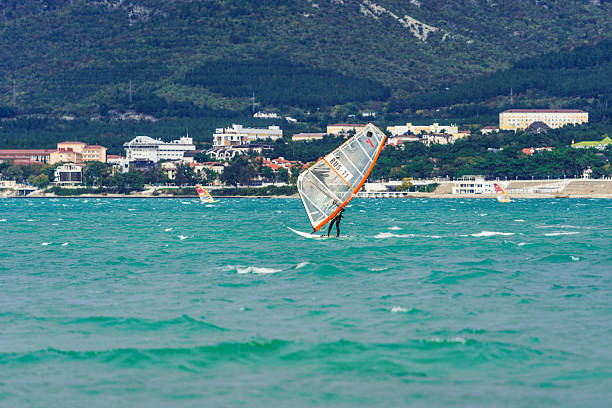 Two people are windsurfing in the ocean. One of them is wearing a red and orange sail