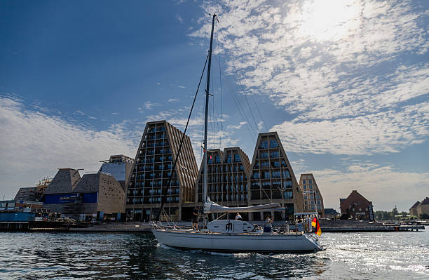 Copenhagen, Denmark-August 14, 2025: White sailing yacht with German flag glides past modern waterfront apartments under blue sky with partial clouds and morning sun.