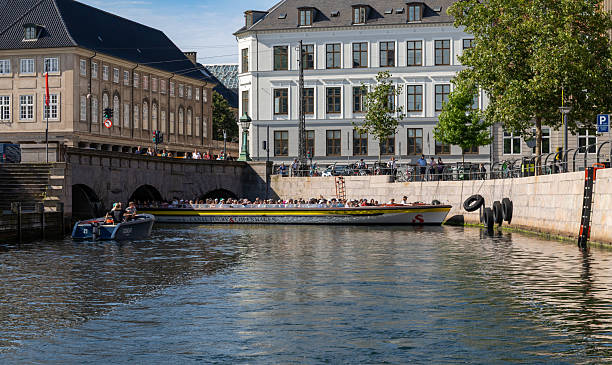 Copenhagen, Denmark-August 14, 2025: Sightseeing boat full of tourists glides under stone canal bridge near old town, surrounded by classic buildings and trees.