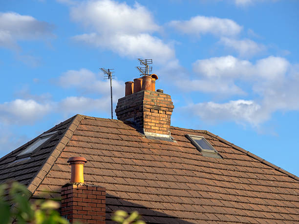 View of a brick chimney on a tiled house rooftop under a clear blue sky with fluffy clouds.