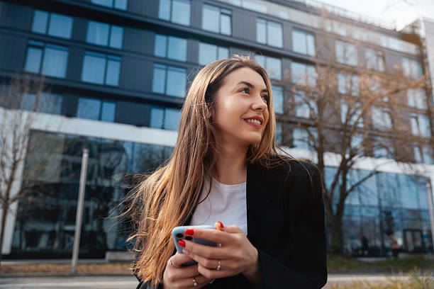 istock Young woman smiling while holding smartphone outdoors in urban setting with modern architecture in background showcasing lifestyle and technology concept 2242068215