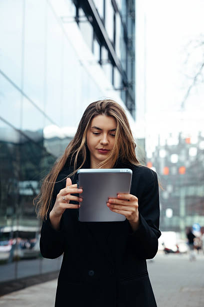 istock Young Woman Using a Tablet on a City Street While Dressed in a Formal Outfit During Daytime Near Modern Buildings and Glass Facades 2242068263
