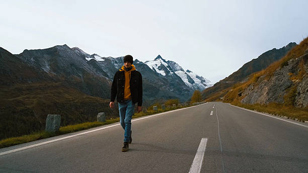 istock Young man walks along an empty road surrounded by majestic mountains in autumn 2242068407