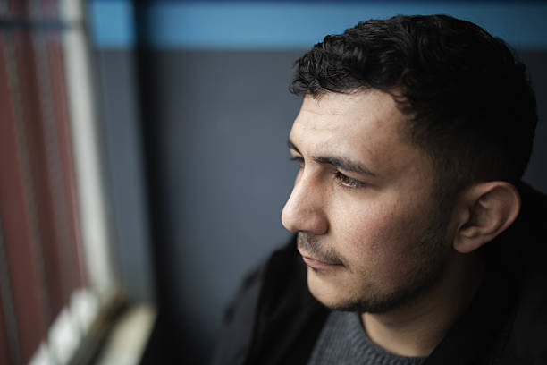A 4K stock photo capturing a young Middle Eastern man gazing thoughtfully through a barred window on a rainy day. His expression combines sadness, pride, and longing as he looks out toward the urban neighborhood below. The atmosphere reflects themes of migration, identity, hope, and melancholy — symbolizing the inner emotions of someone possibly far from home. Perfect for illustrating concepts such as refugee life, urban solitude, nostalgia, or human resilience.