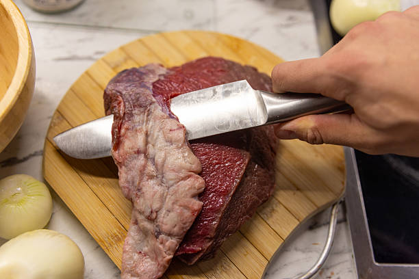 A hand holds a sharp knife, slicing raw beef on a wooden cutting board in a kitchen. Crunch of meat and prep vibes convey cooking, dinner, and meal preparation.