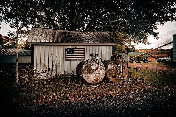 A weathered white farm shed stands beneath a large tree, featuring a faded American flag mounted on its wall and surrounded by old rusted fuel tanks. The scene captures the texture and quiet authenticity of rural America — a timeless portrait of work, endurance, and pride in the countryside.This image embodies themes of Americana, agriculture, history, and resilience, perfect for editorial use or projects exploring U.S. rural identity and nostalgia.