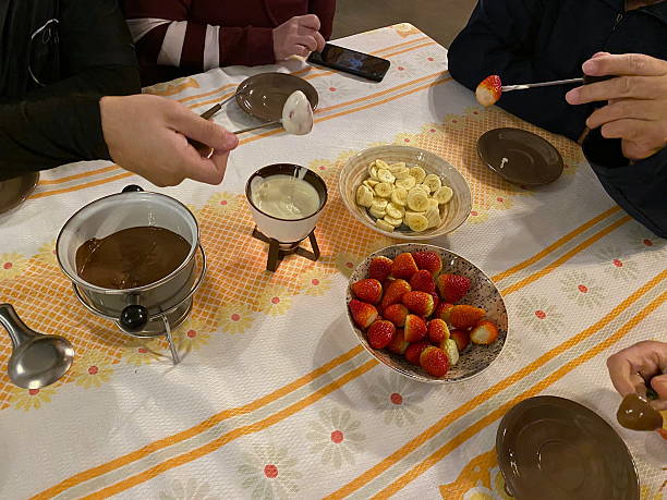A top view of a dining table in a cozy setting, where several people share a delicious chocolate fondue. The center of the table is dominated by a small metal cauldron on a stovetop, filled with melted, creamy chocolate, next to a smaller bowl of white chocolate. There are bowls of fruit for dipping: a large one with fresh, vibrant red strawberries and another with banana slices. Hands hold fondue forks, dipping strawberries into the syrup, highlighting the social and fun aspect of the meal. The table is covered with a white tablecloth decorated with stripes and flowers in shades of yellow and orange.