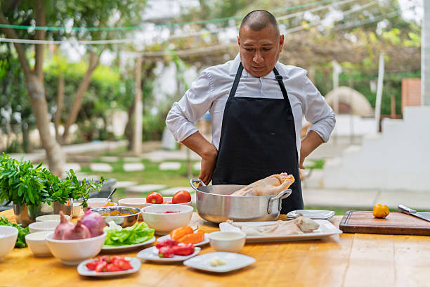 Chef wearing an apron preparing a suckling pig with fresh vegetables and herbs on a wooden table in an outdoor setting