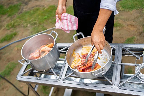 Person using tongs to cook fresh meat in a large pot on a portable gas stove outdoors, preparing a delicious meal