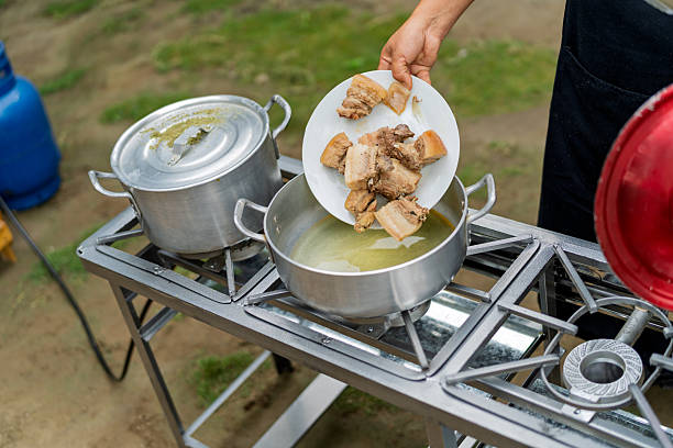 Person's hand adding pieces of fresh pork meat from a white plate into a pot on an outdoor gas stove