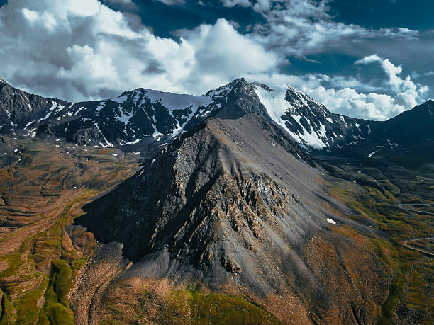 Aerial view of a sharp, rocky mountain ridge leading to snow-capped peaks, overlooking a winding valley with red and green terrain