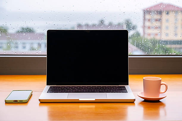 istock Laptop with blank screen, smartphone and coffee cup on wooden table. Remote work setup on rainy day. 2242105726
