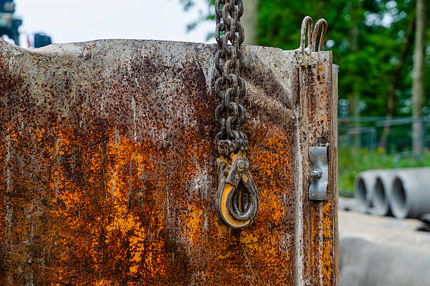 A rusty crane rigging displays a heavy chain hook used for lifting construction materials. The background shows trees and construction equipment in use.