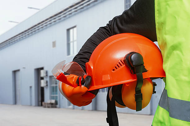 istock Construction worker in high visibility vest holding orange hard hat with headlamp and safety goggles at industrial site. Personal protective equipment for workplace safety and construction projects. 2242114669