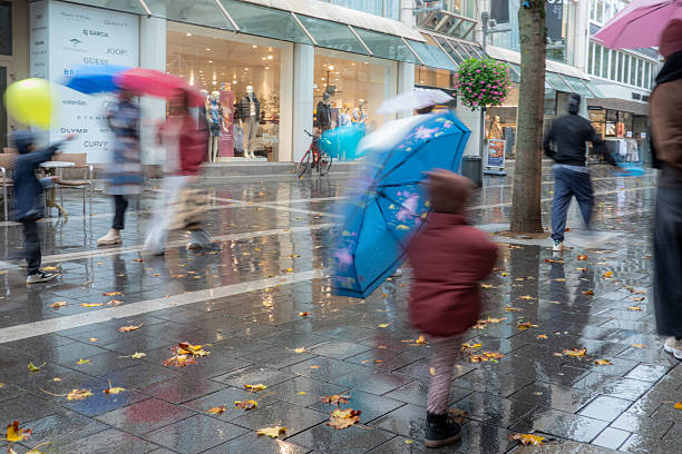 Urban street scene during autumn rain featuring people in blurred motion with umbrellas and shopping reflections