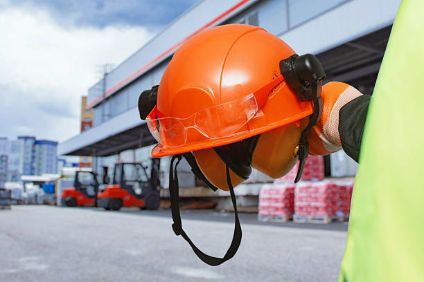 istock Orange construction helmet with earmuffs and safety goggles held by a worker in high-visibility gear at a warehouse site. Industrial safety, personal protective equipment in logistics. 2242116148