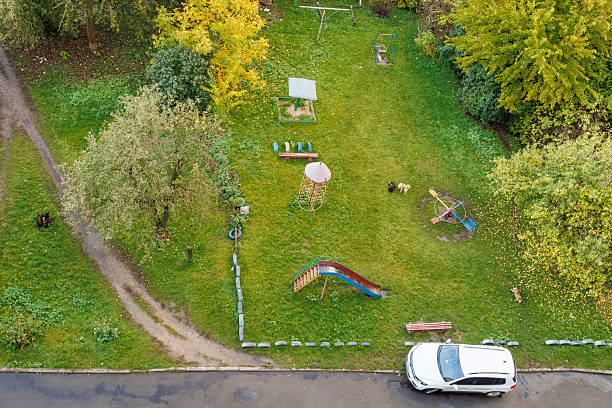 Aerial view of an old playground with a slide, swing, and merry-go-round surrounded by green grass, trees, and a parked white car.