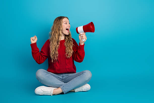 istock Pretty young woman holding a red megaphone while seated, expressing happiness on bright blue background indoors 2242124813