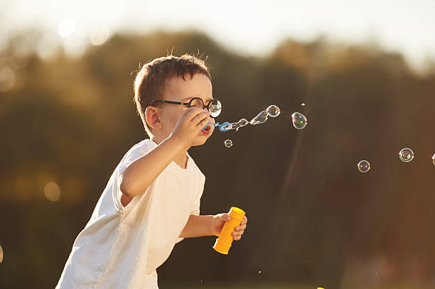 Standing and blowing bubbles, focused. Boy is having fun on the field under sunlight at daytime.