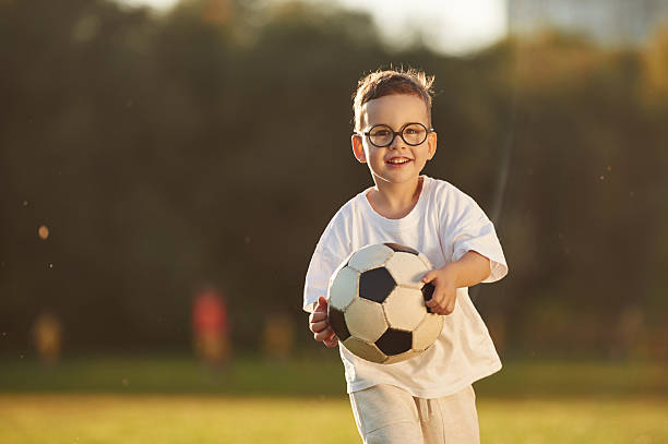 Holding soccer ball, front view. Boy is having fun on the field under sunlight at daytime.