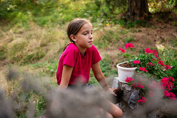 Child girl is sitting in the yard surrounded by flowers during summer day