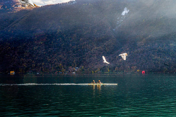 A mesmerizing view of a kayak paddling across Lake Lugano with two women  winter in the canton of Ticino: a healthy lifestyle in the southern canton of Switzerland. A small patch of snow on the mountain. Seagulls in the air (backlit by the sun's rays). Christmas is coming soon.